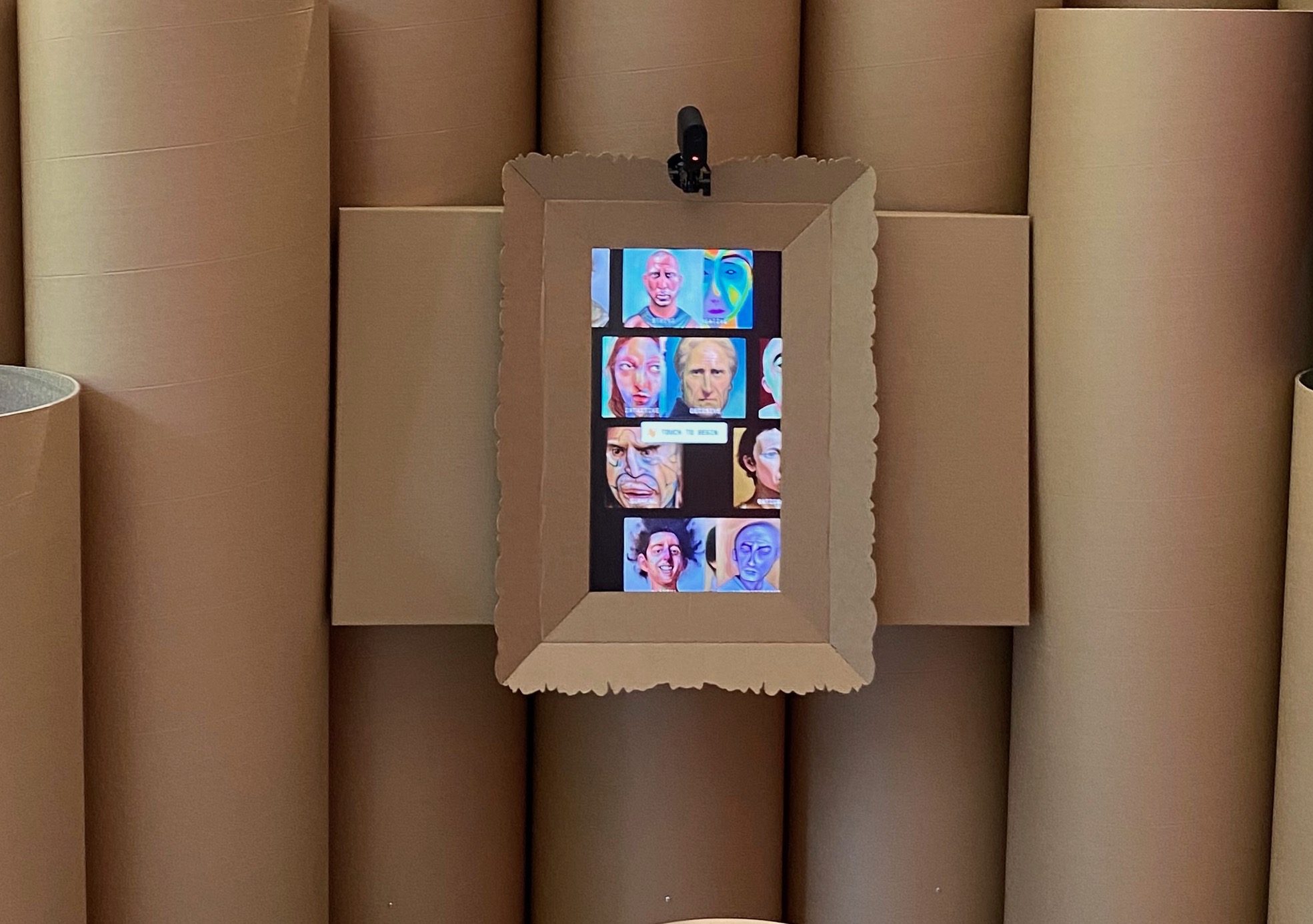 A close up of self portrait installed on brown cardboard poles and surrounded by a brown cardboard frame. the touchscreen shows a screensaver of different generative AI images and a webcam