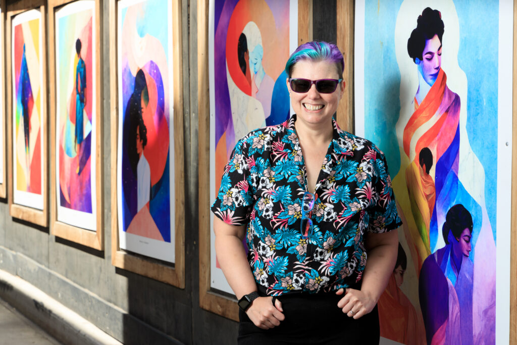 AI artist J. Rosenbaum standing in front of some of their works, they are wearing a bright shirt covered in skulls and tropical plants which matches their hair in shades of turquoise and pink. they are wearing sunglasses and smiling.