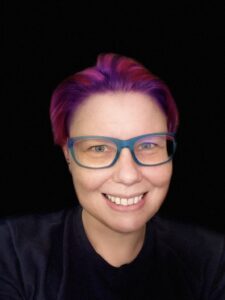 a headshot of Melbourne AI artist J. Rosenbaum. They have short red and purple hair (subject to change) blue and pink glasses and pale skin with freckles.
